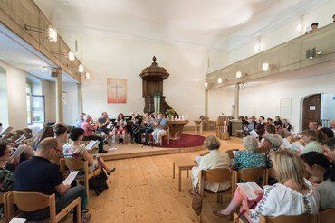 Innenraum einer Kirche mit Menschen, die in einem Halbkreis sitzen und einem Altar zugewandt sind.