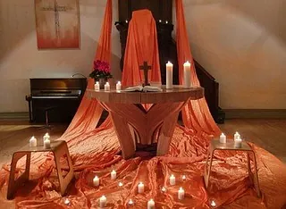 Altar, geschmückt mit orangefarbenem Stoff, Kreuz und vielen Kerzen.